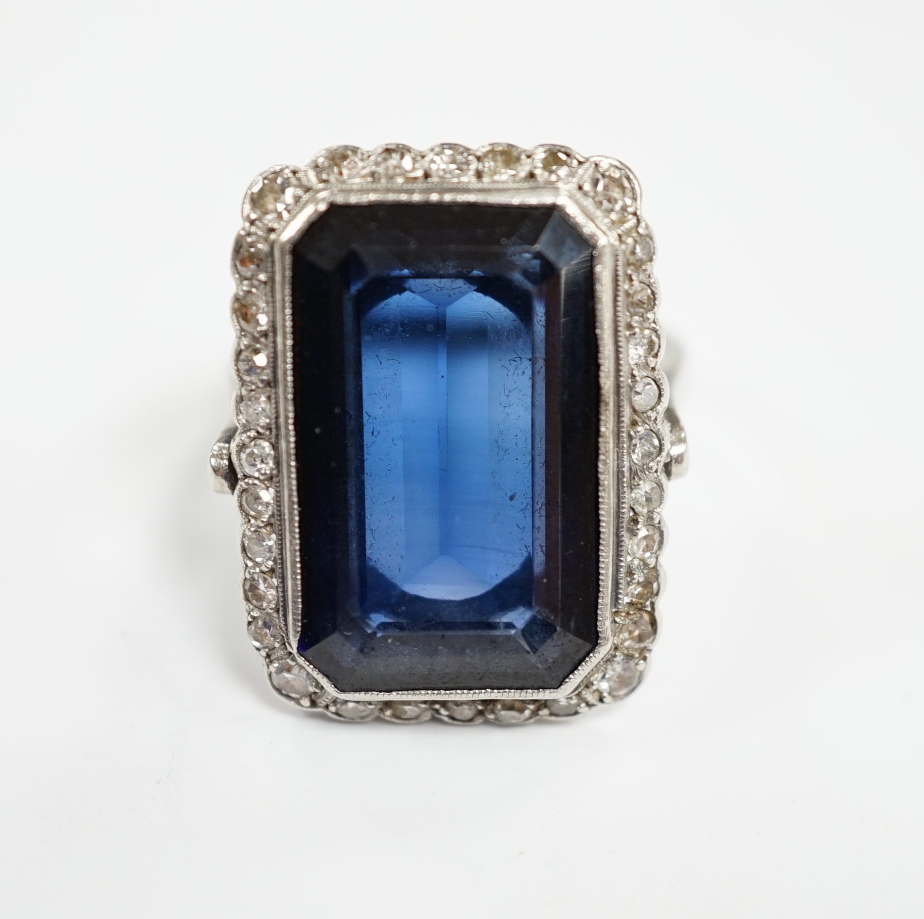 A 1920's white metal and single stone emerald cut synthetic sapphire set dress ring, with millegrain set diamond chip border, size N, gross weight 10 grams.